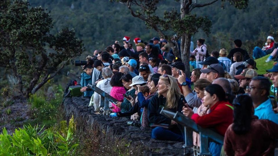 Rows of people stand behind a low rock wall protecting them from a cliff edge.