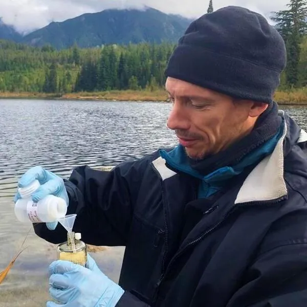 A man standing near a small body of water collecting samples in a plastic container.