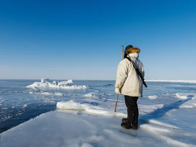 A person wearing full Arctic gear holds a harpoon, standing on the sea ice and looking out into the frozen ocean