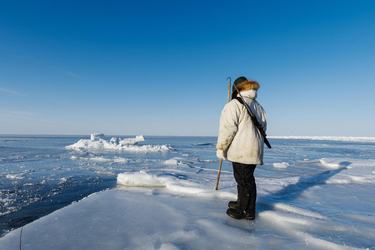 A person wearing full Arctic gear holds a harpoon, standing on the sea ice and looking out into the frozen ocean