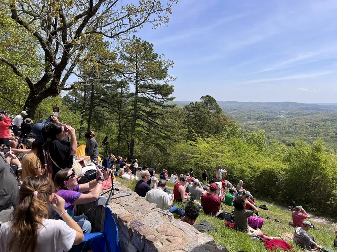 A crowd of people sit in an open field with a forested landscape in the background.