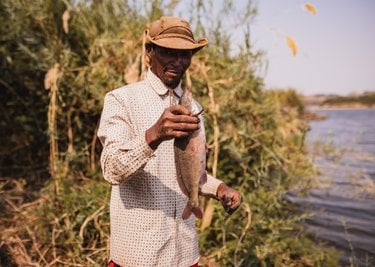 A man in a hat stands holding a fish by a river bank