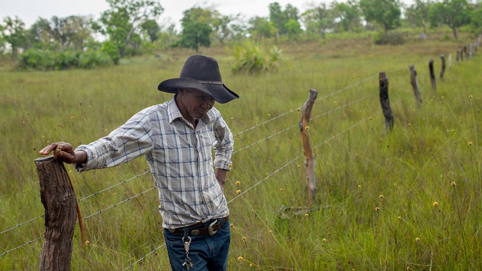 A man leans against a fence post in a field of tall grass, wearing a large hat that shades his face