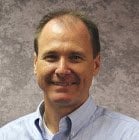 A middle-aged white man smiles in front of a grey background. He is wearing a light blue button down.