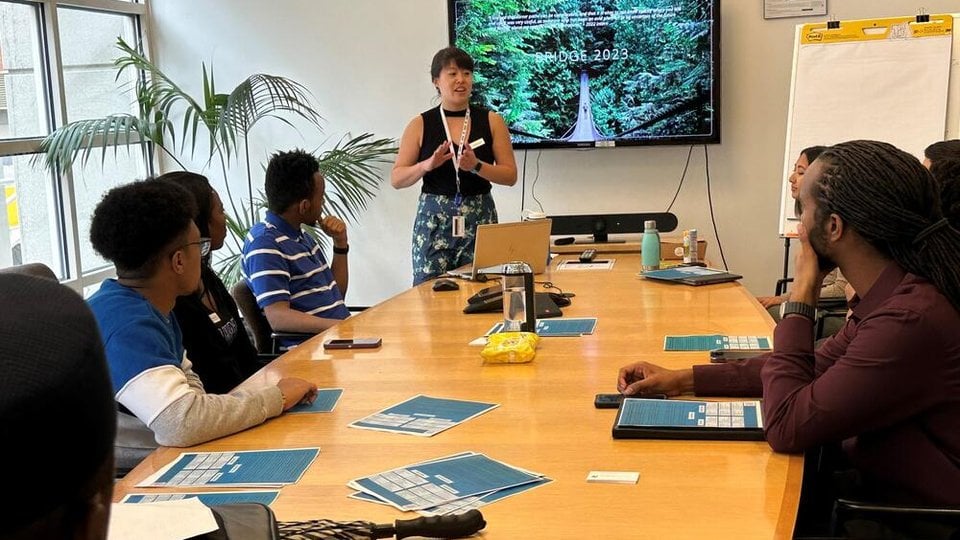 A woman stands in front of a TV monitor with a group of people seated around a table in a conference room.