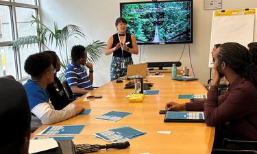 A woman stands in front of a TV monitor with a group of people seated around a table in a conference room.