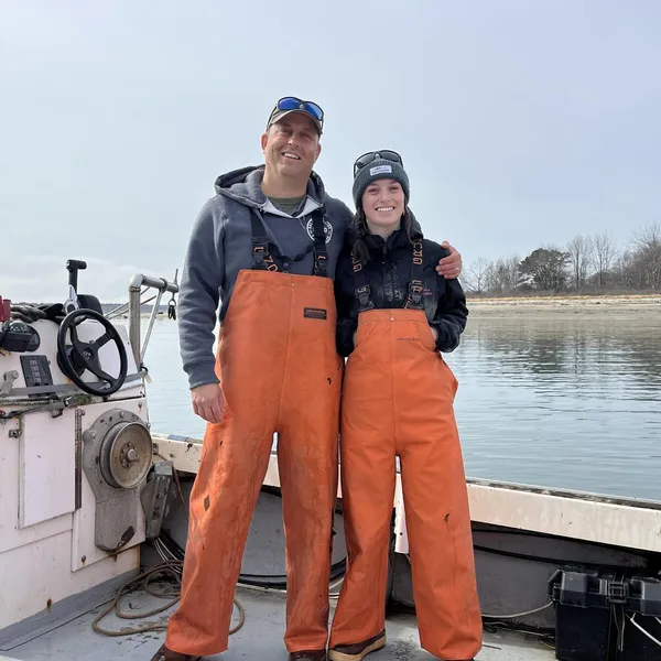 A man and woman in orange fishing gear on a fishing boat on water.
