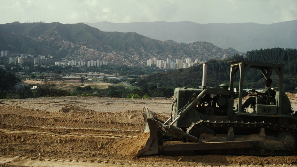 Expanding and mountainous city of Caracas in the background. In the foreground, a bulldozer flattens the ground.