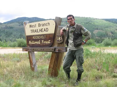 A man wears outdoor gear and stands in front of a trailhead sign in the Roosevelt National Forest.