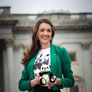 A woman with long hair, a green sweater, and a black and white tshirt with the WWF panda logo, holds a stuffed panda in front of a government building.
