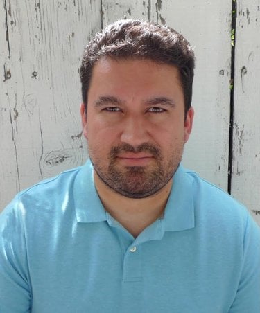 A white man with dark, short-cropped wavy hear and short beard. He is wearing a blue shirt.