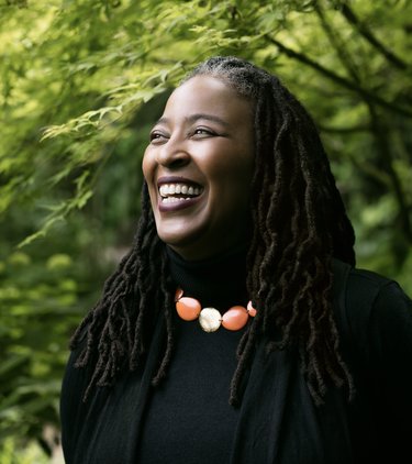 Smiling headshot of Camille Dungy. Her locs fall below the shoulder, and she is wearing all black, a large red and gold necklace. She looks off to the right. In the background are trees.