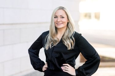 A middle-aged white woman with long blonde hair smiles with her hands confidently on her waist. She is wearing an all-black outfit.
