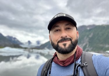 Craig Santos Perez smiles. In the background are mountains and a glacial lake.