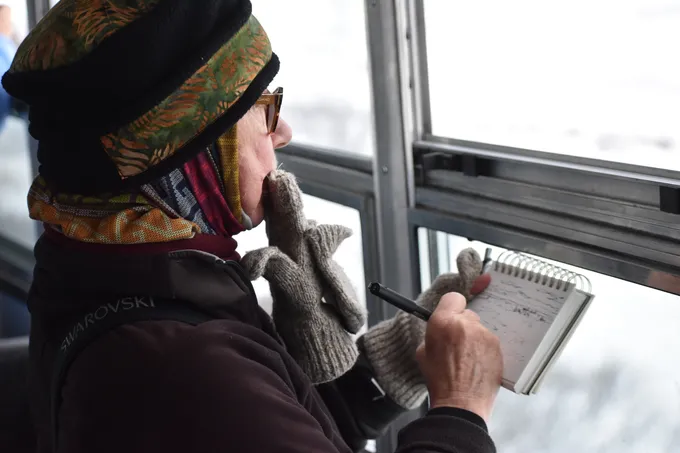 A woman with a sketchpad gazing out a window, her glove in her mouth