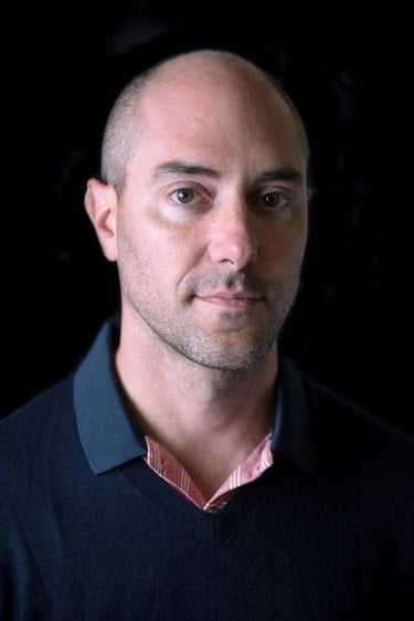 A bald, white man against a dark background gently smiles. He is wearing a dark navy button down sweater.