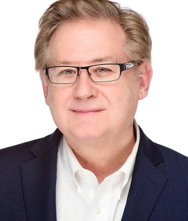 An middle-aged white man with dark framed glasses in front of a white background wears a dark blue suit and gently smiles.