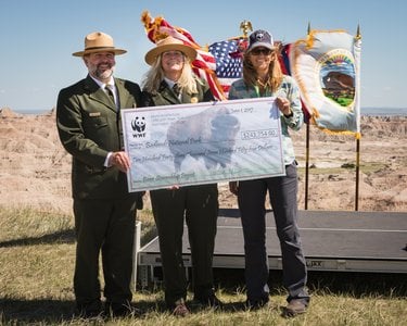 Two park rangers in uniforms stand in front of flags and hold an oversized check with a third person in a WWF hat