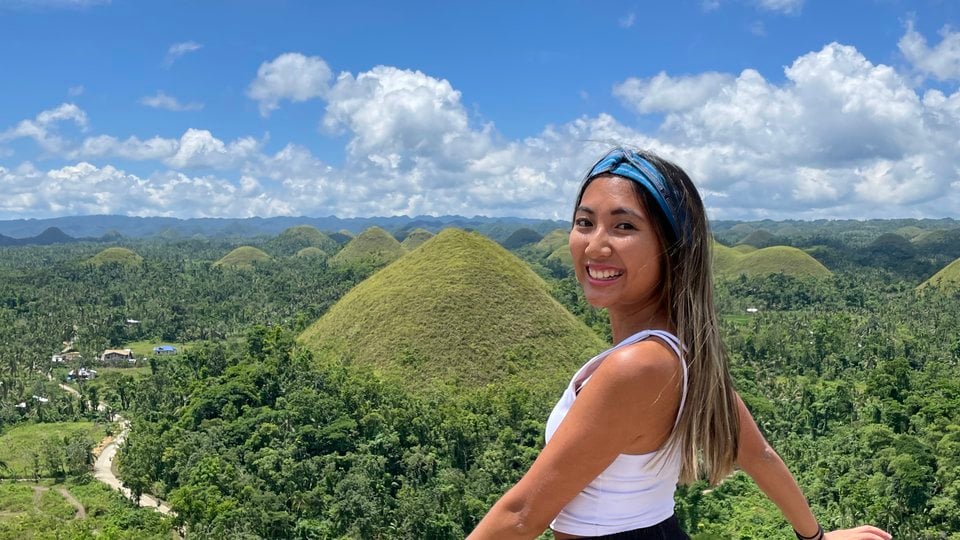 Francesca Edralin stands above a scenic view of forests and mountains with a cloudy blue sky looking back at camera