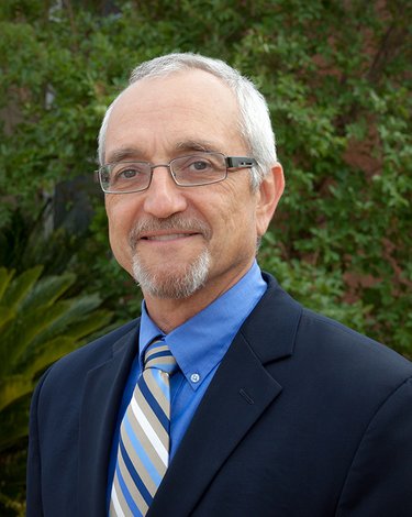 An Italian man with greying hair wears a navy blue suit and blue shirt. He is wearing glasses and smiles into the camera.