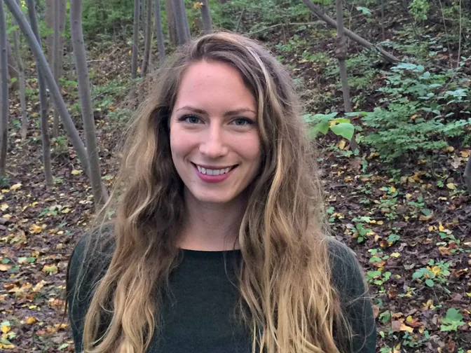 Giavanna Grein wears a black shirt and smiles at the camera in a wooded area