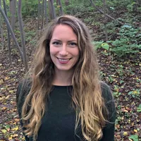 Giavanna Grein wears a black shirt and smiles at the camera in a wooded area
