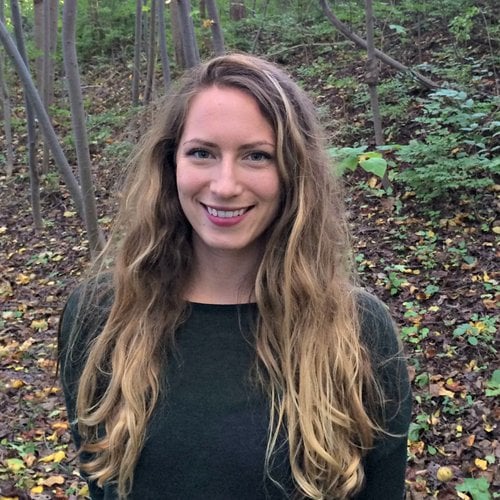 Giavanna Grein wears a black shirt and smiles at the camera in a wooded area