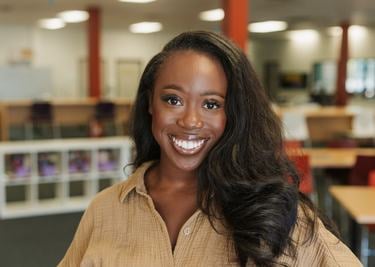 Loretta Lyken stands in a library and wears a tan buttondown shirt and has long black hair and smiles at the camera