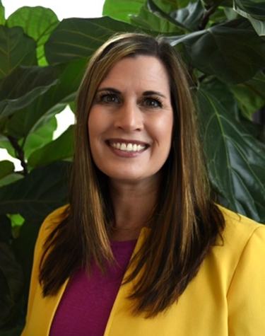 Gerin Hennebaul has long dark hair and wears a yellow jacket and pink shirt and smiles at the camera in front of greenery