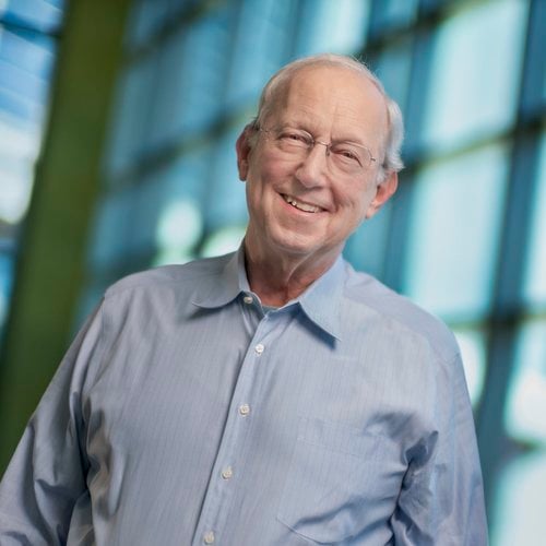John Sall smiles at the camera with large windows seen in the background behind him