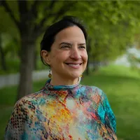 Josefina smiles looking off towards the left. She is wearing a colorful turtleneck sweater. Behind her is a lush park.