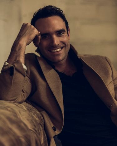 Joshua smiles, leaning on a suede couch with his forehead resting on his knuckles. He is wearing a tan coat and black shirt.