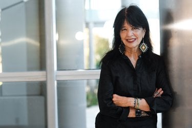 Joy leans on a metal pillar and smiles. She is wearing a black tunic and large beaded earrings and has her arms crossed in front of her.