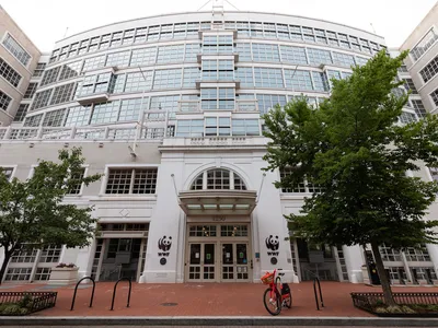 The facade of a white building with a curved front, many windows, a brick sidewalk and trees.