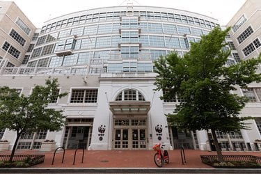 The facade of a white building with a curved front, many windows, a brick sidewalk and trees.