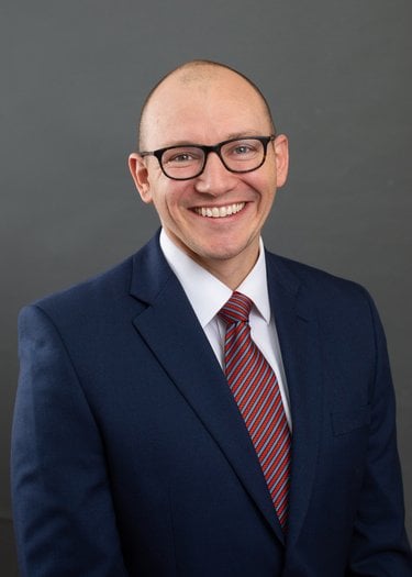 A bald white man with dark framed glasses smiles. He is wearing a blue suit, white shirt, and patterned red tie.