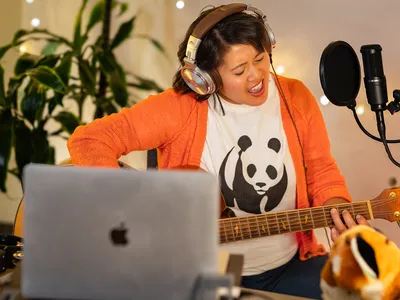 A musician wearing a WWF shirt plays a guitar and sings into a mic, a computer nearby