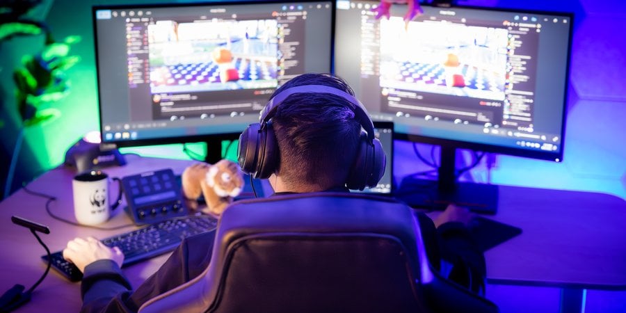 Looking at a video gamer from behind. He's in a dark room lit up with LED lights and has two monitors.
