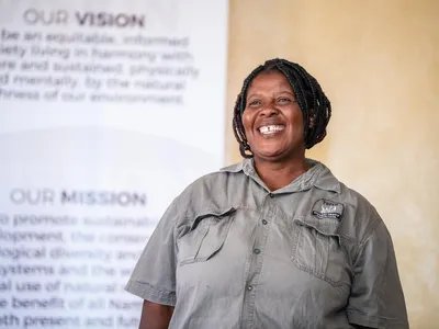 Constansia Somses, Game Guard at Torra Conservancy attends a female Rhino Rangers meeting at Uibasen Twyfelfontein Conservancy head office, Namibia, stands in front of a poster in a gray shirt and smiles at the camera