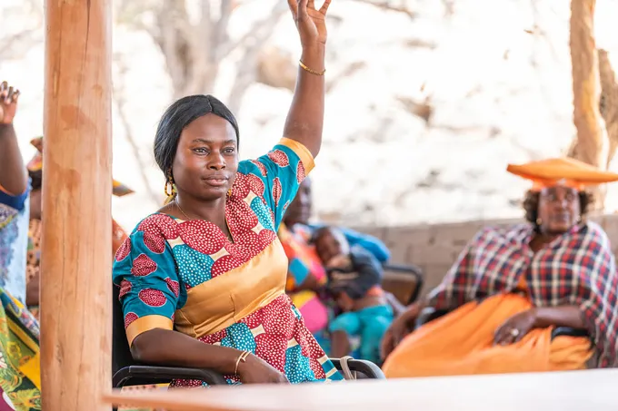 Tjikaa Katjirumbu wears a red, blue, and gold outfit and raises a hand with other people in brightly colored clothing in the background