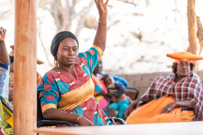 Tjikaa Katjirumbu wears a red, blue, and gold outfit and raises a hand with other people in brightly colored clothing in the background