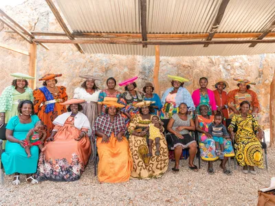 Members of Woman for Conservation sit and stand in two rows wearing brilliantly colored clothing and hats under a wood structure