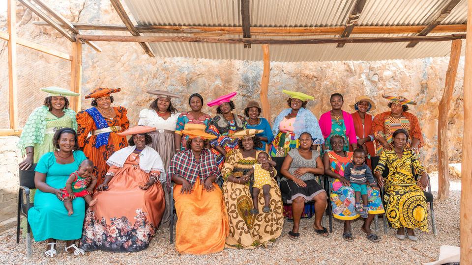 Members of Woman for Conservation sit and stand in two rows wearing brilliantly colored clothing and hats under a wood structure