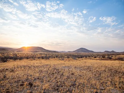 View from Etendeka Mountain Camp in Namibia shows sparse brown plants and mountains in the distance with the sun peeking over the top of one and blue sky with scattered clouds