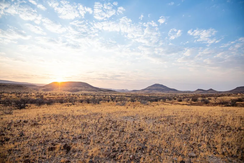 View from Etendeka Mountain Camp in Namibia shows sparse brown plants and mountains in the distance with the sun peeking over the top of one and blue sky with scattered clouds