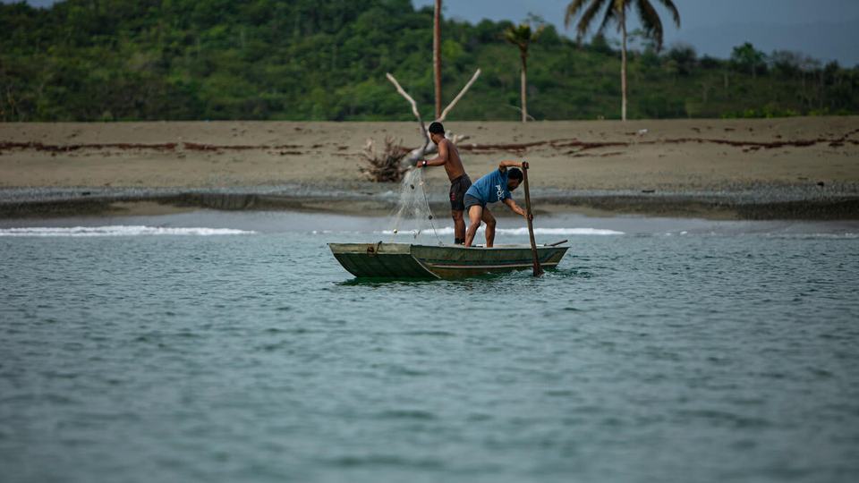Two fishers in a small wooden boat manage nets close to shore with a beach and forest-covered hill in the background