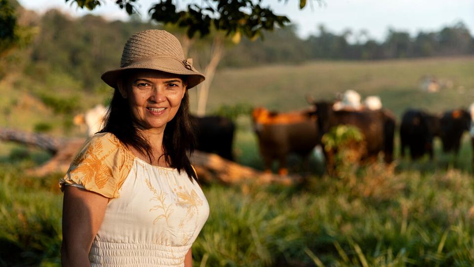 Veronica Cardozo wears a hat and a white shirt with yellow sleeves and smiles at the camera with her cattle in the background