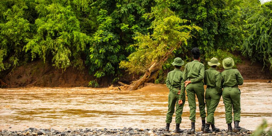 Four rangers in green uniforms with green hats stand on the bank of a brown river looking across to lush forest