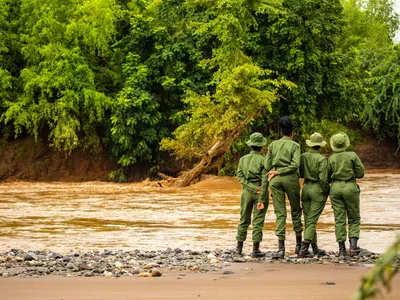 Four rangers in green uniforms with green hats stand on the bank of a brown river looking across to lush forest