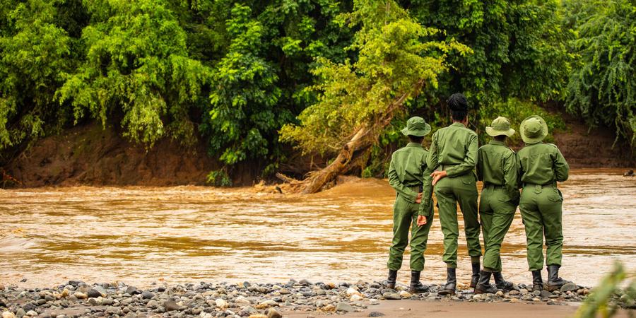 Four rangers in green uniforms with green hats stand on the bank of a brown river looking across to lush forest